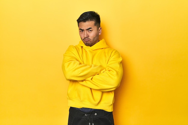 Photo d'un homme asiatique audacieux en sweat à capuche jaune sur un studio jaune, fronçant les sourcils de mécontentement, garde les bras croisés