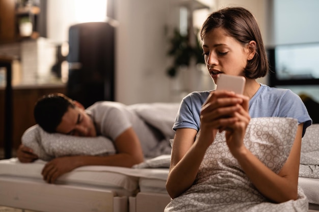 Photo gratuite une jeune femme envoie secrètement des messages texte sur son téléphone intelligent et trompe son petit ami qui dort sur le lit
